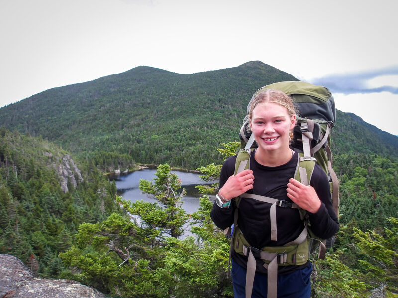 A young woman with blonde hair stands smiling on a mountain top, wearing a large hiking backpack. Behind her, a lush green forest stretches out, revealing a small lake nestled among the trees. In the distance, a larger mountain rises, partially obscured by clouds. The foreground features rocky terrain and evergreen foliage.